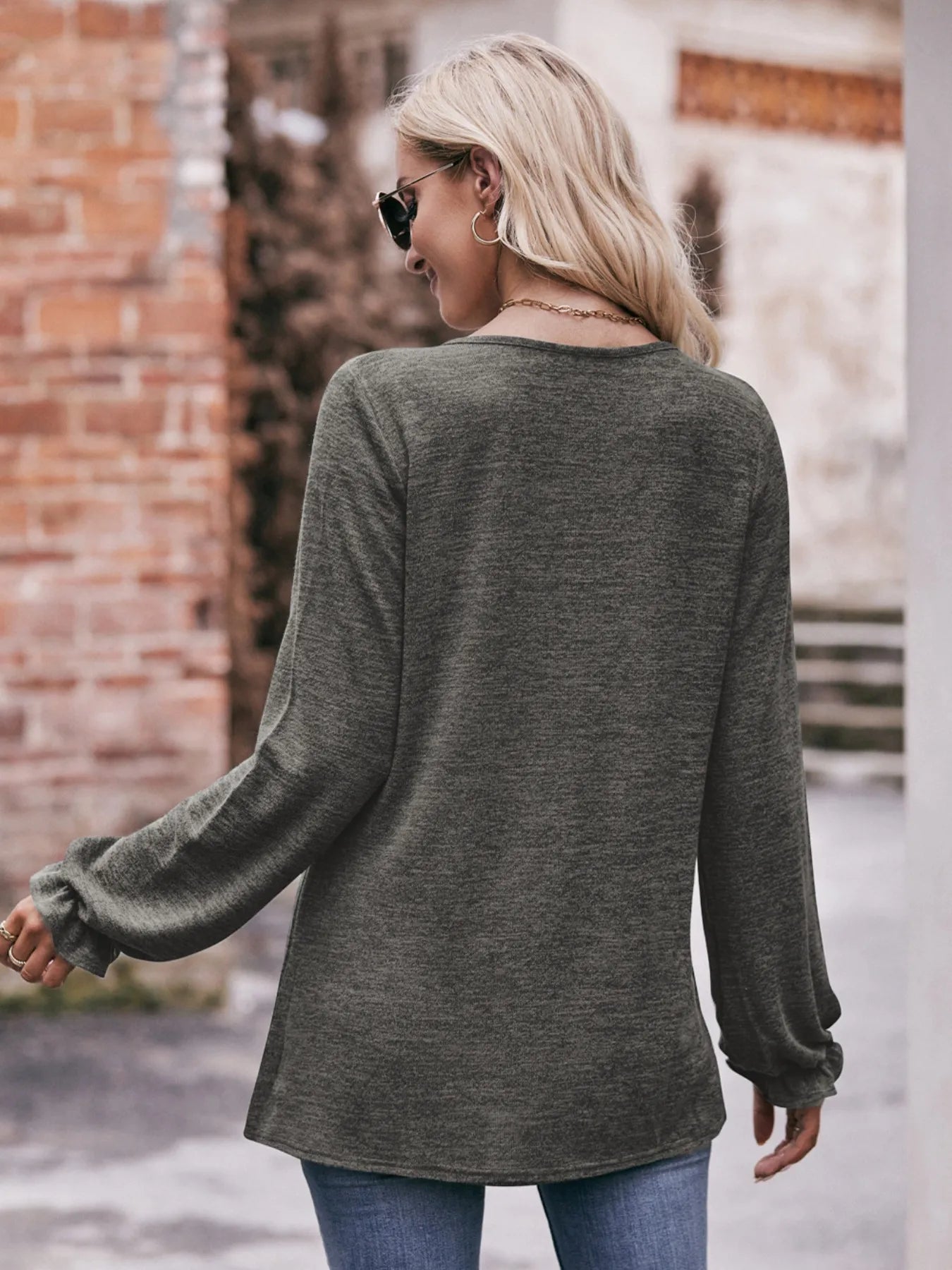 Woman wearing a green long-sleeve top and blue jeans, standing against a brick wall.