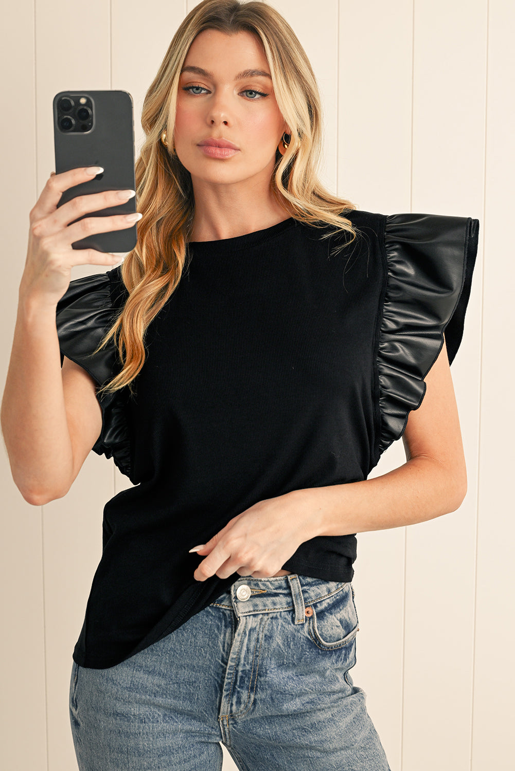 Woman wearing a black top with ruffled sleeves and blue jeans, taking a selfie.