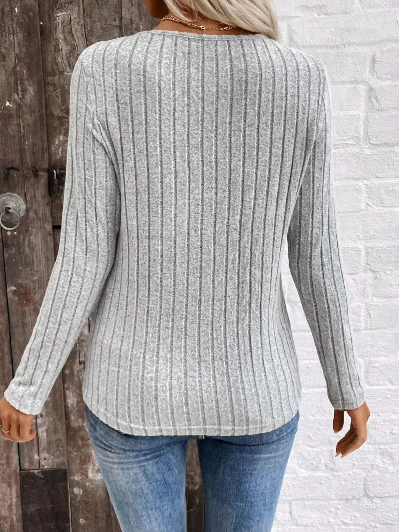 Person wearing a gray ribbed sweater and blue jeans against a white brick wall.