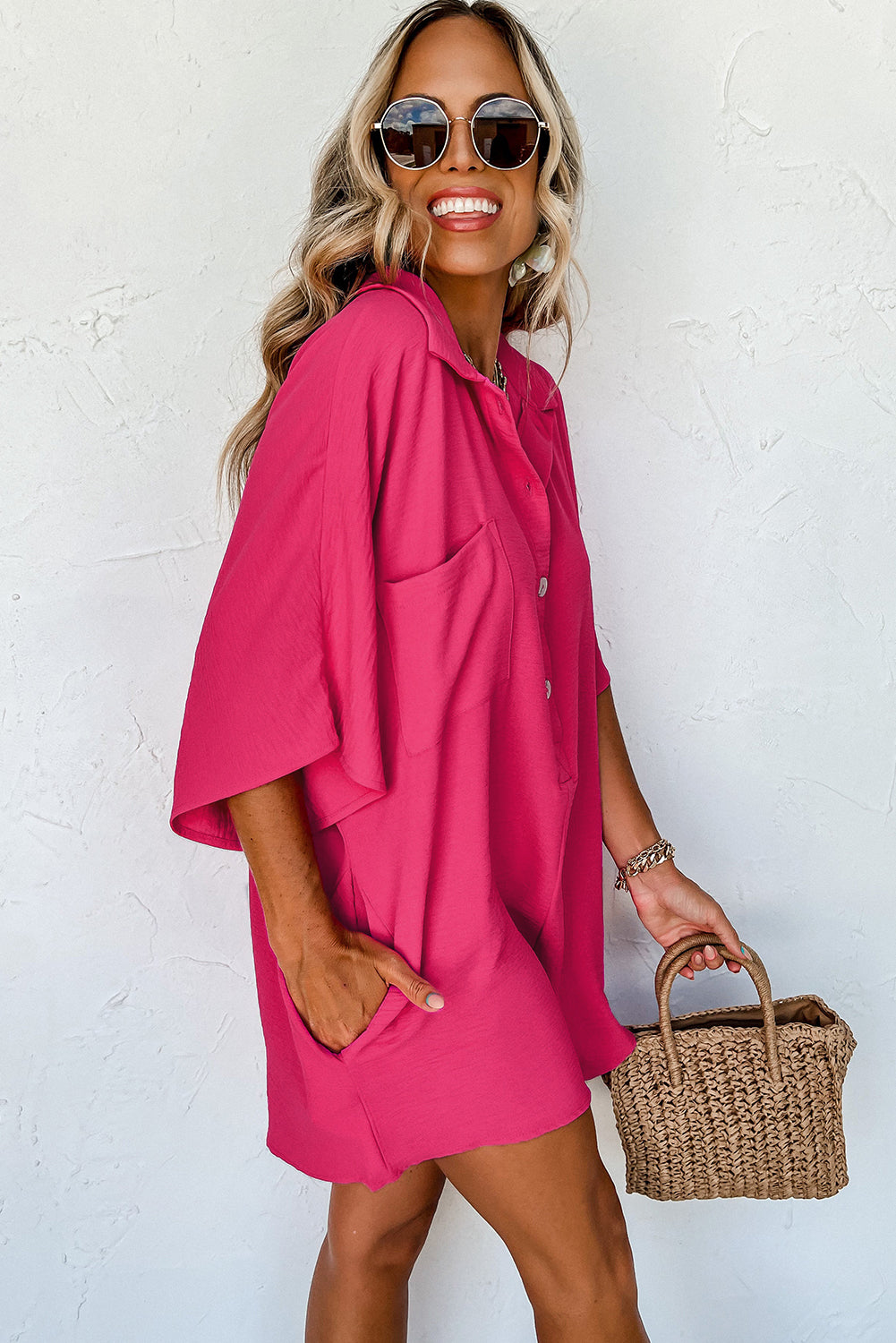 Woman wearing a bright pink dress with sunglasses and a straw bag against a white background