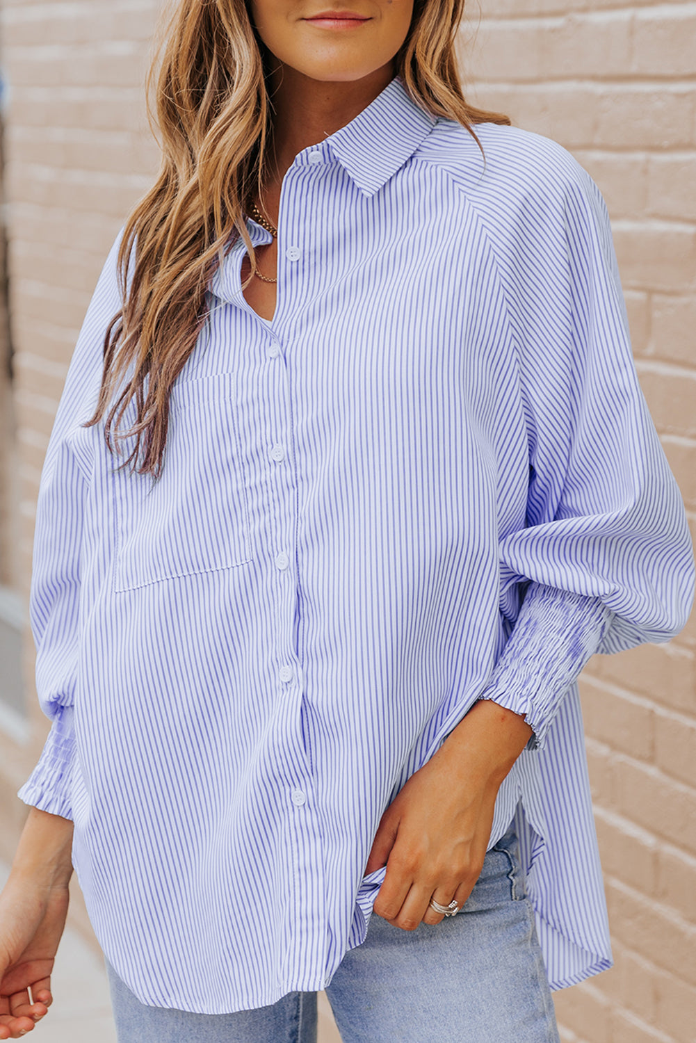 Woman wearing a blue and white striped shirt with rolled-up sleeves against a beige wall.
