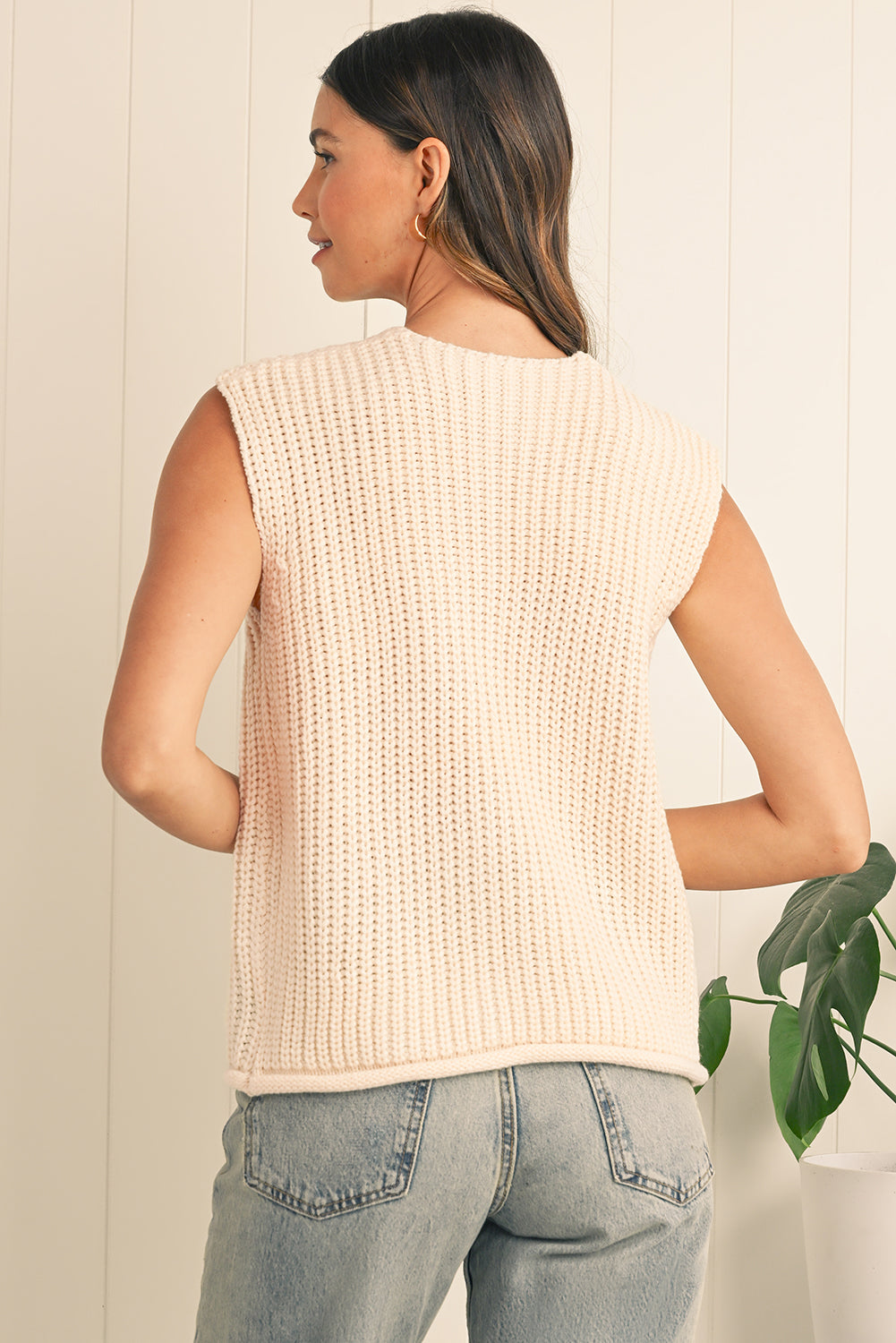 Woman wearing a beige knitted vest and jeans against a neutral background