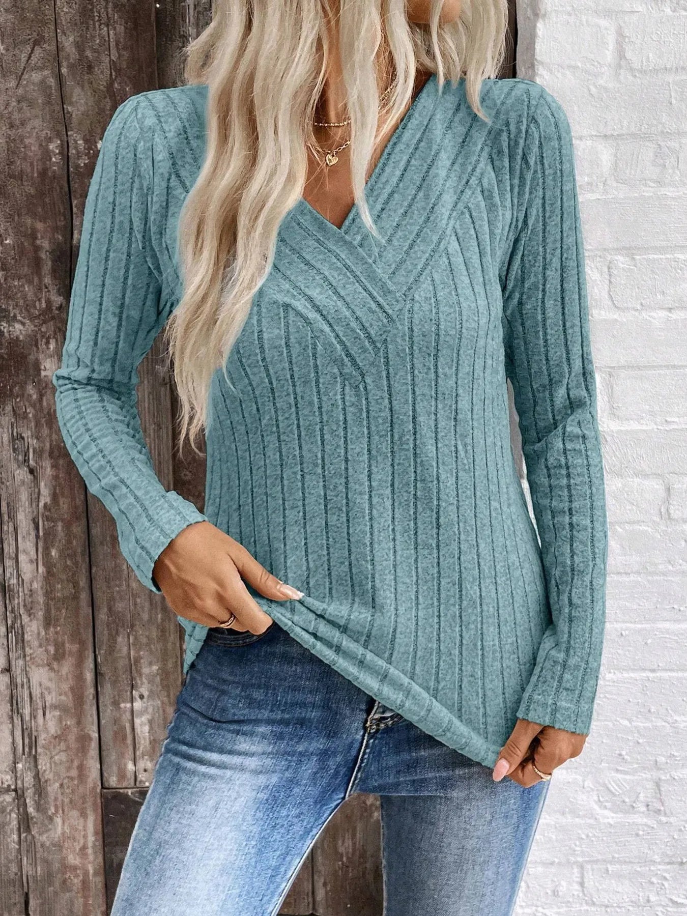 Woman wearing a teal ribbed sweater and blue jeans against a wooden and brick wall.
