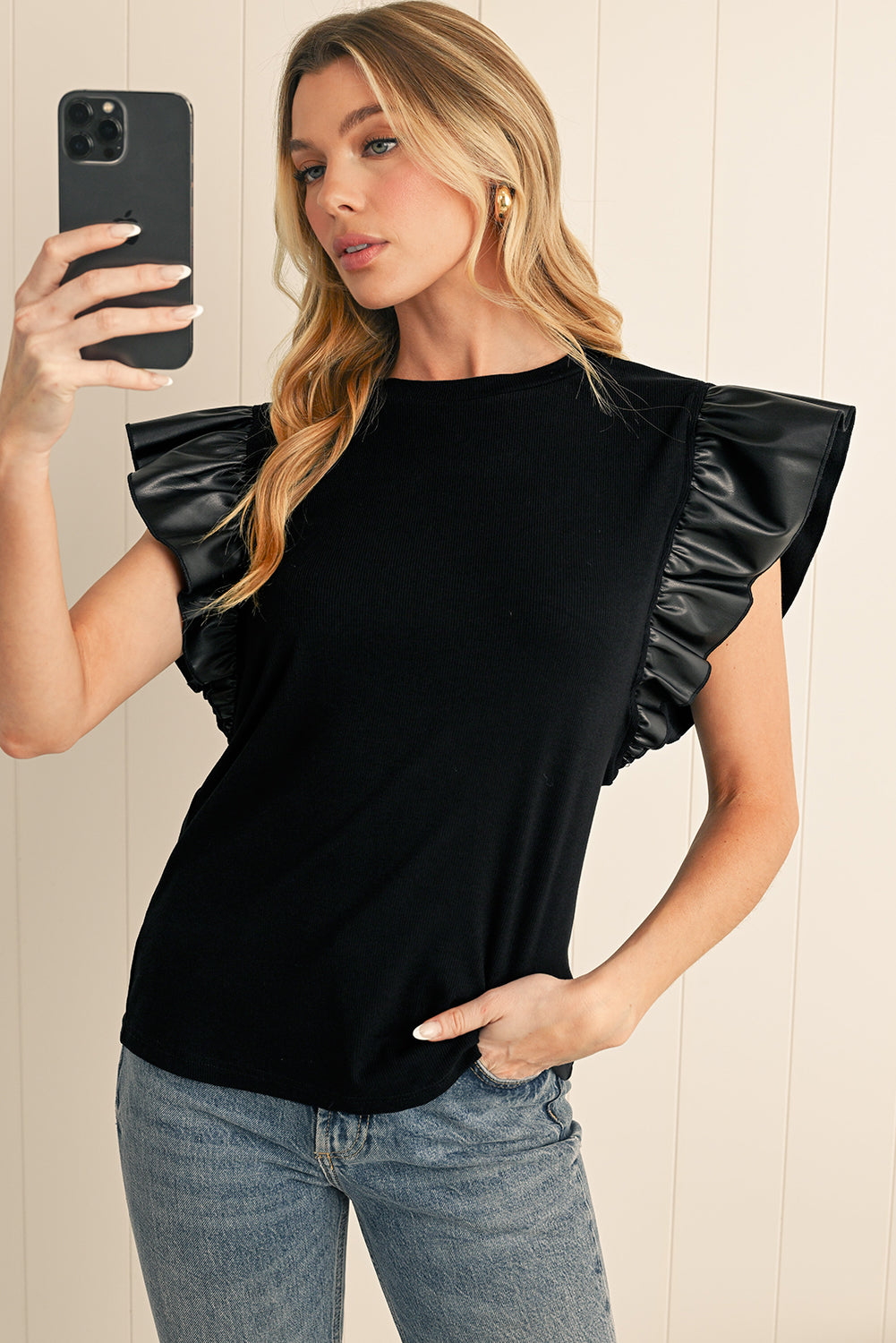 Woman wearing a black ruffled sleeve top and blue jeans, taking a selfie.