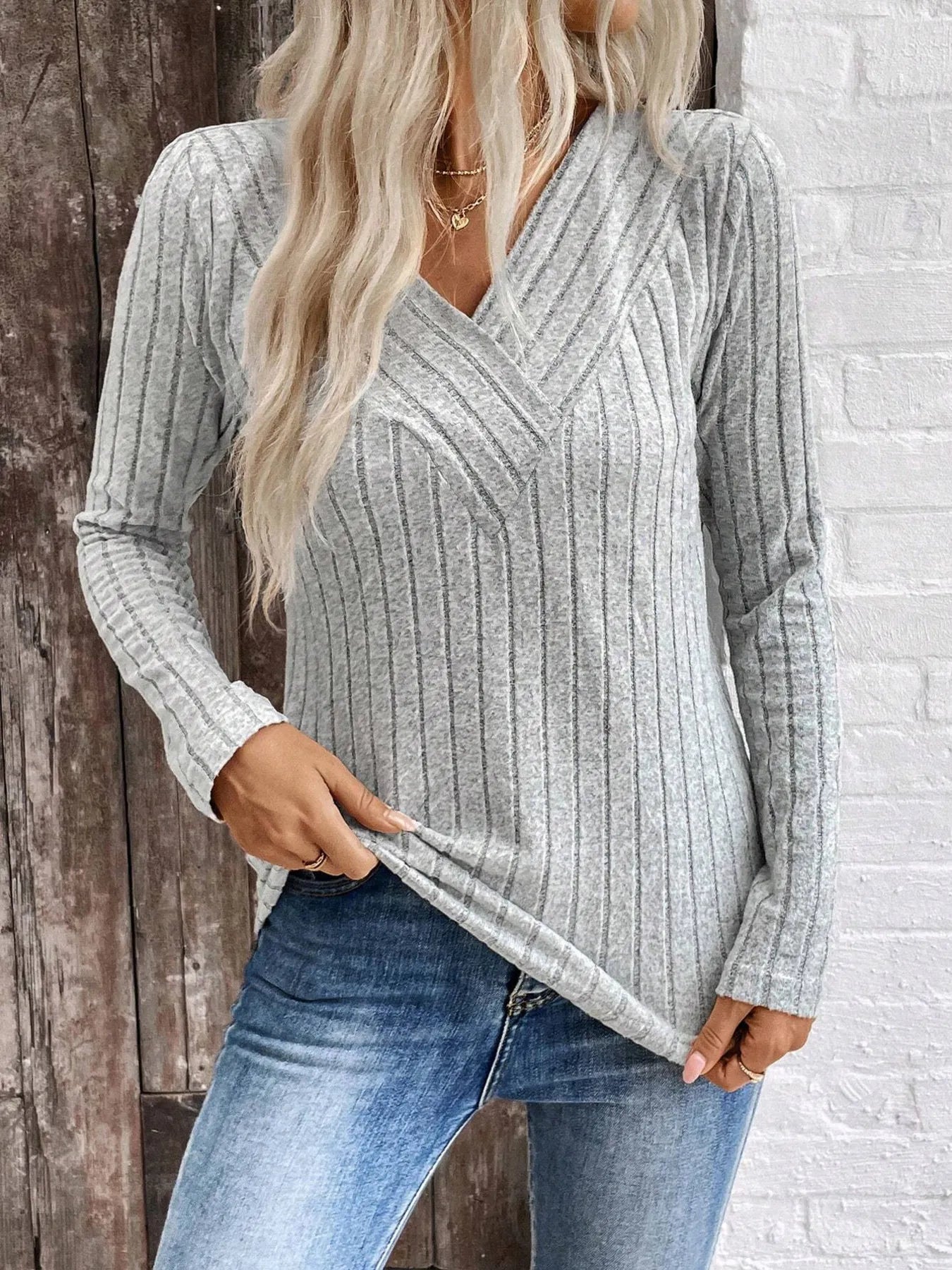 Person wearing a gray ribbed sweater and blue jeans against a wooden and brick wall.
