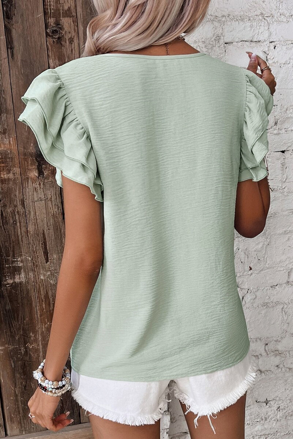 Person wearing a light green top with ruffled sleeves against a wooden and brick wall.