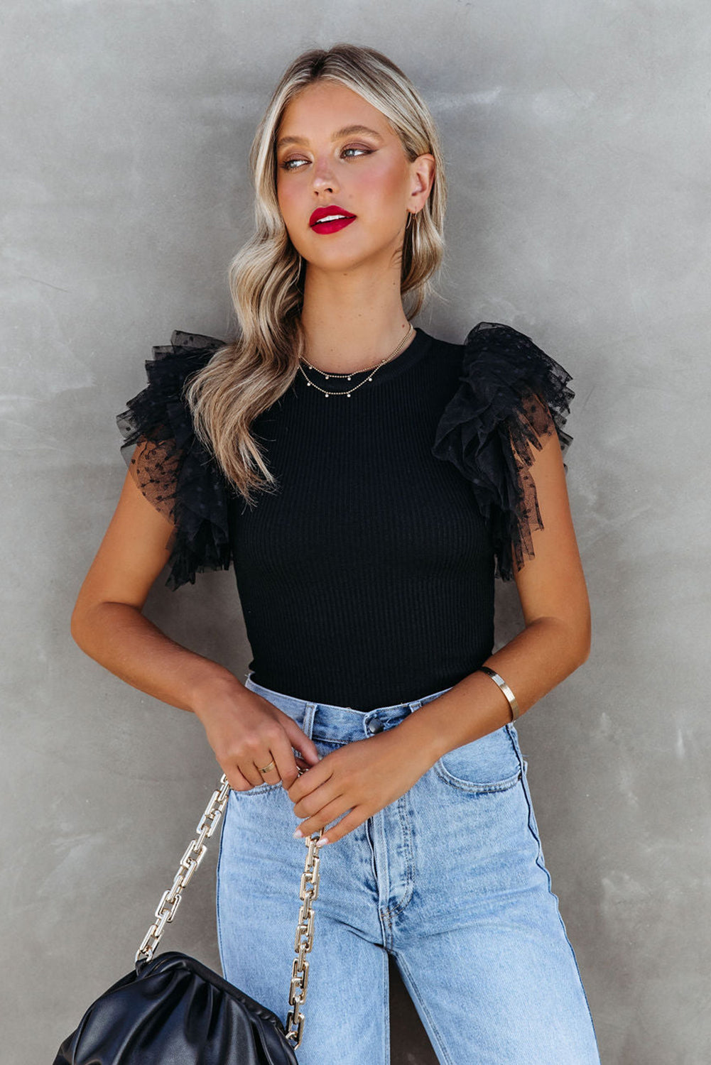 Woman wearing a black top with ruffled sleeves and blue jeans against a gray background