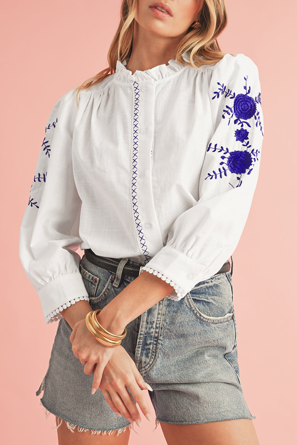 White blouse with blue floral embroidery worn by a person on a pink background