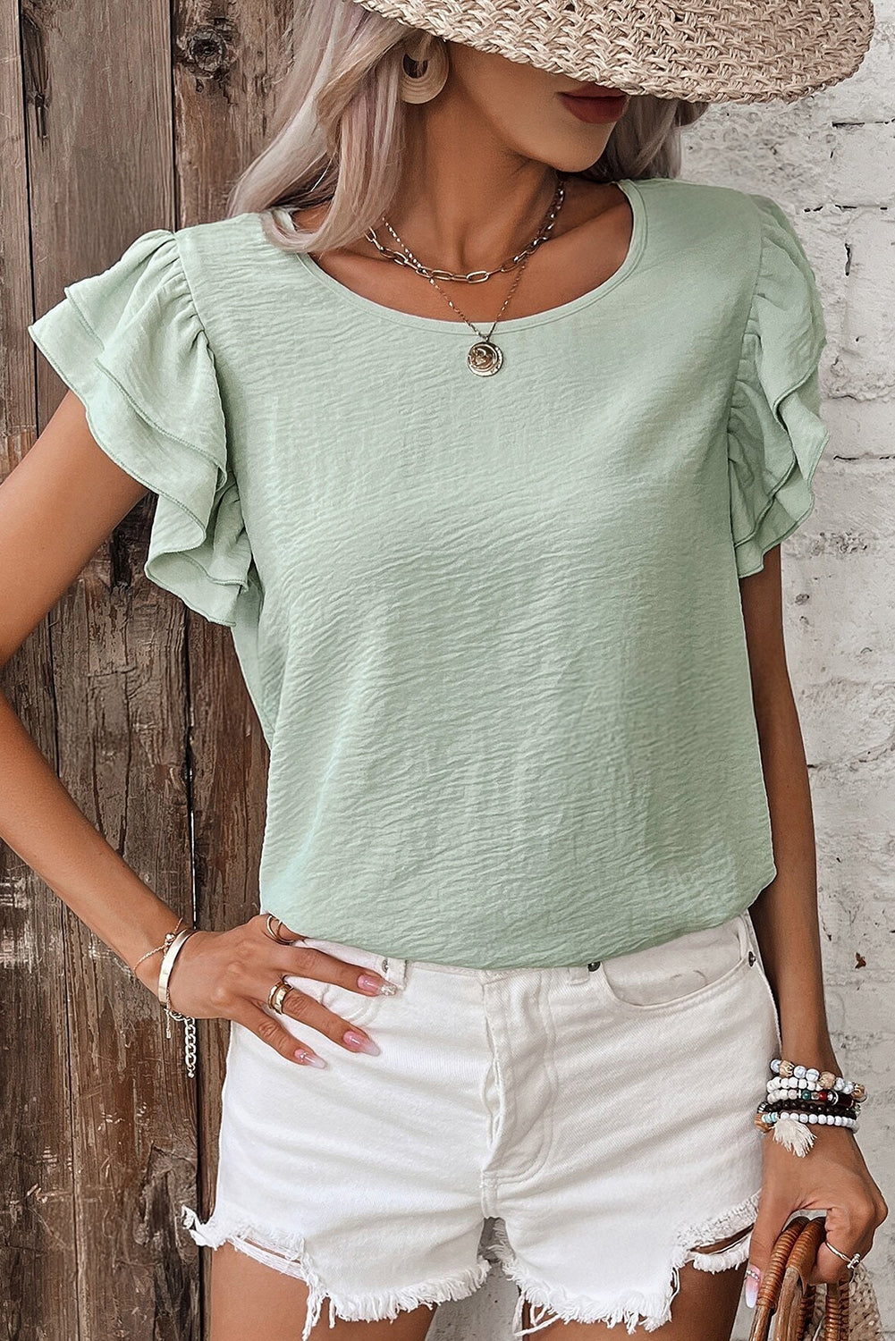 Woman wearing a light green top with ruffled sleeves and white shorts against a wooden and brick wall.