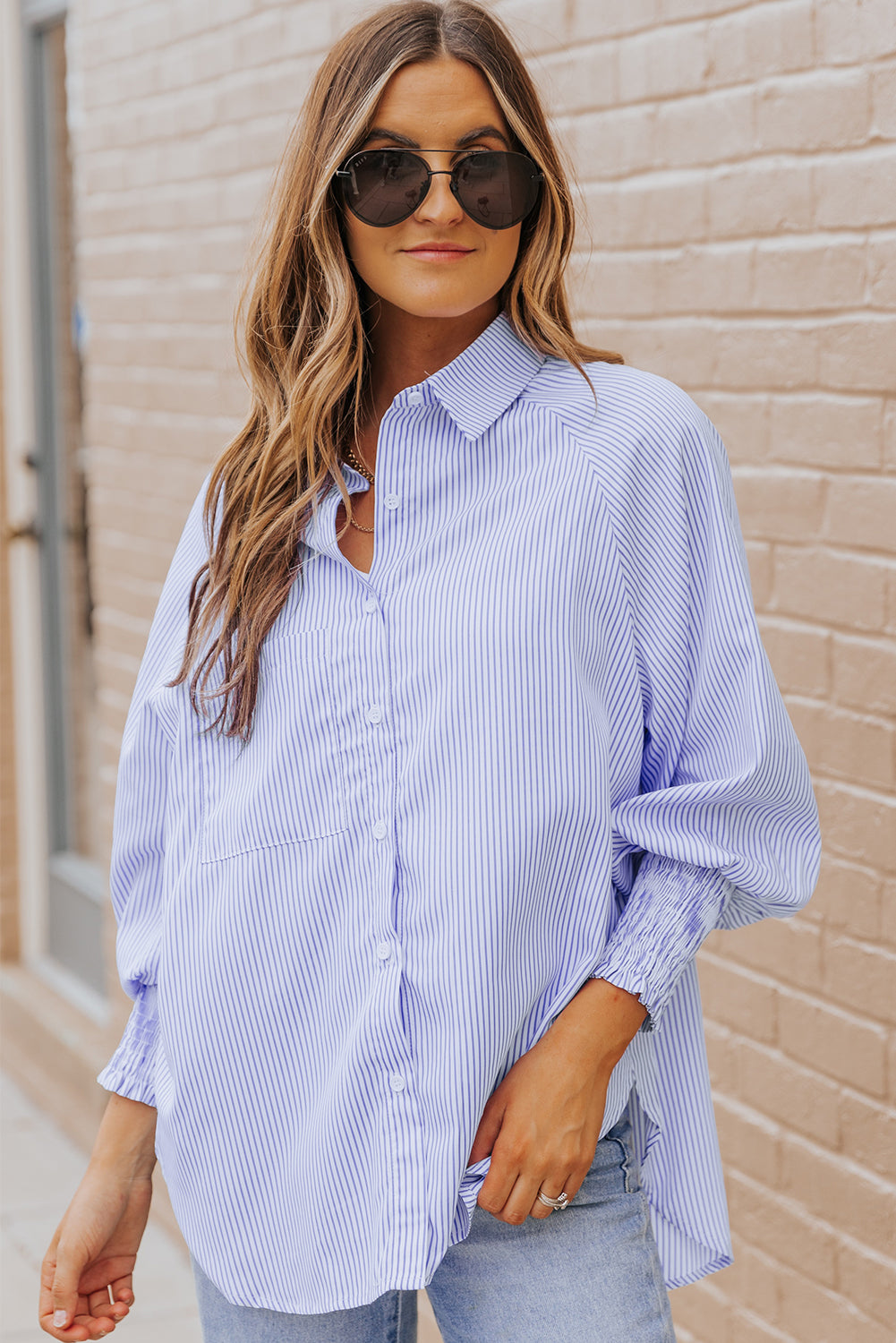 Woman wearing a blue striped shirt and sunglasses against a beige wall.