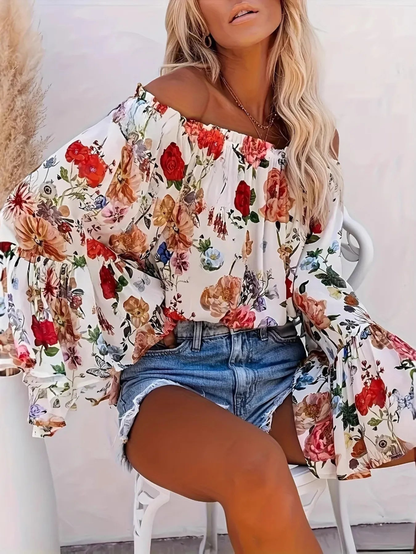 Woman wearing a floral off-shoulder top and denim shorts sitting on a white chair.