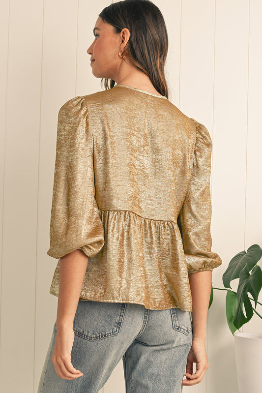 Woman wearing a gold metallic blouse and blue jeans against a white wall.