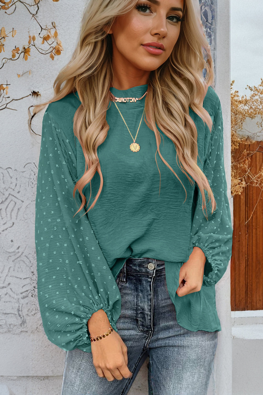 Woman wearing a green long-sleeve top with a subtle pattern, standing against a decorative wall.