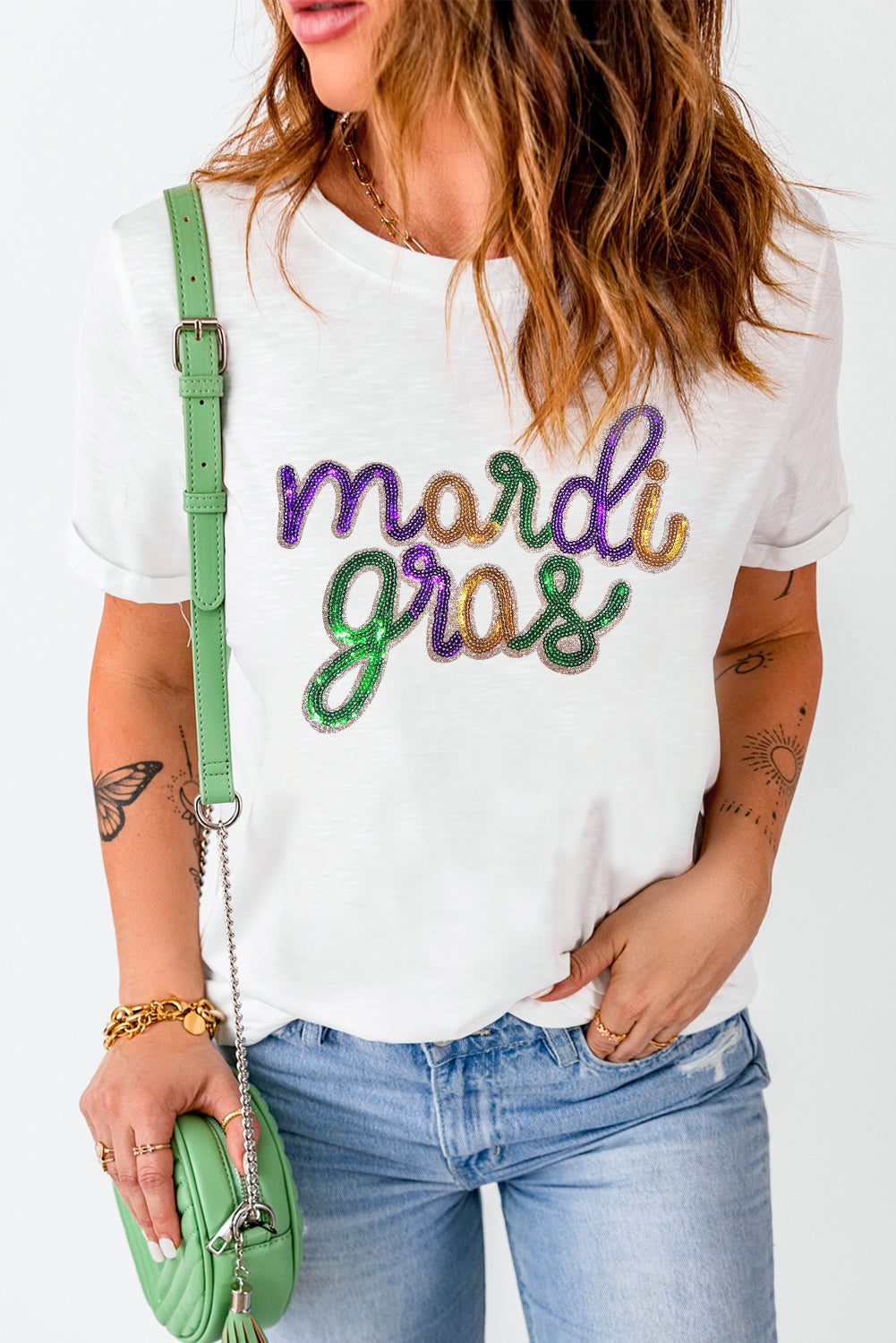 Person wearing a white t-shirt with 'mardi gras' text, holding a green purse.