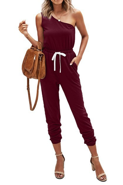 Woman wearing a burgundy one-shoulder jumpsuit with a brown backpack on a white background