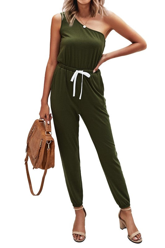 Woman wearing an olive green one-shoulder jumpsuit with a brown bag on a white background