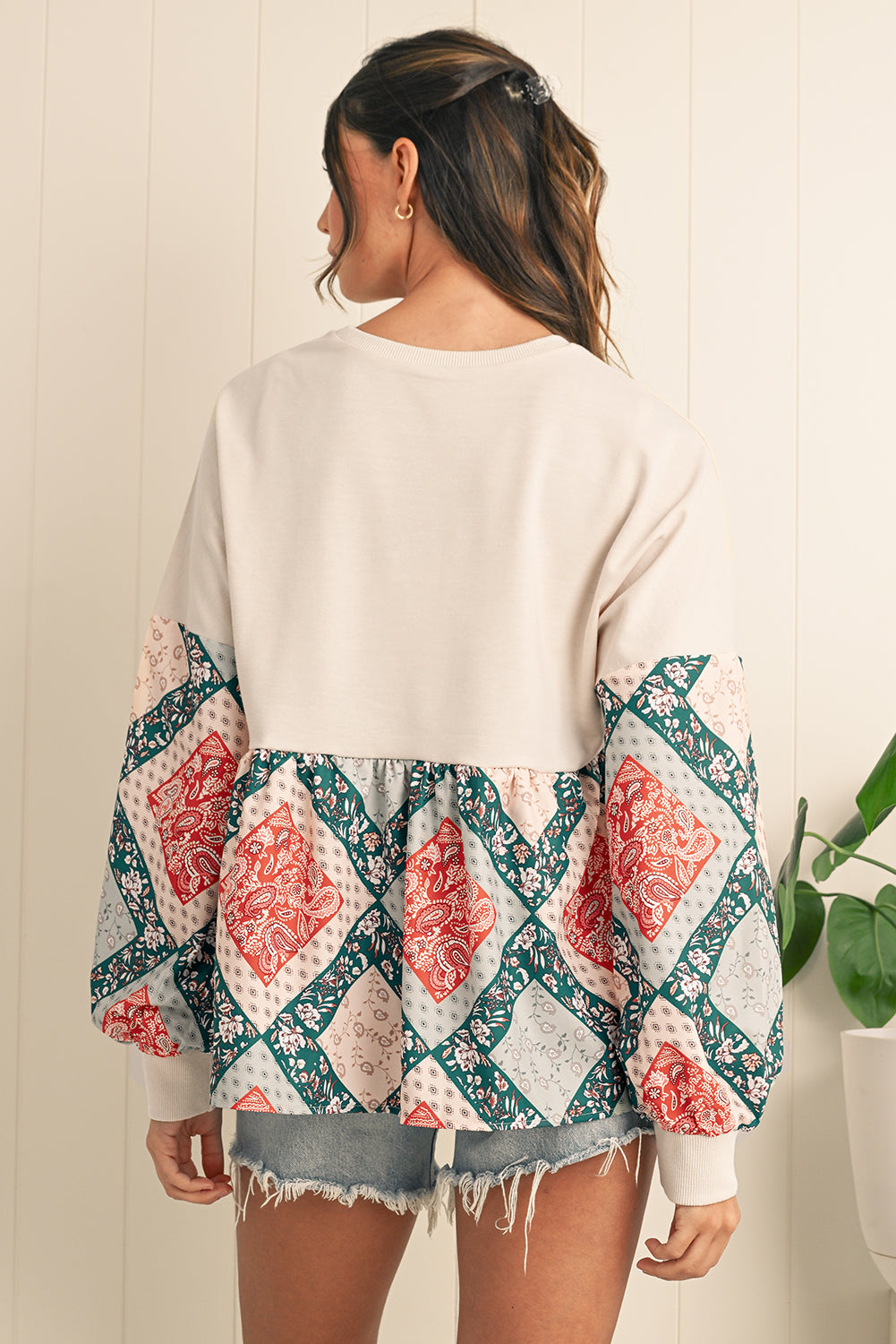 Woman wearing a white sweater with a colorful patterned kimono-style top over it, standing against a light wooden panel background.