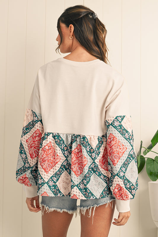 Woman wearing a white sweater with a colorful patterned kimono-style top over it, standing against a light wooden panel background.