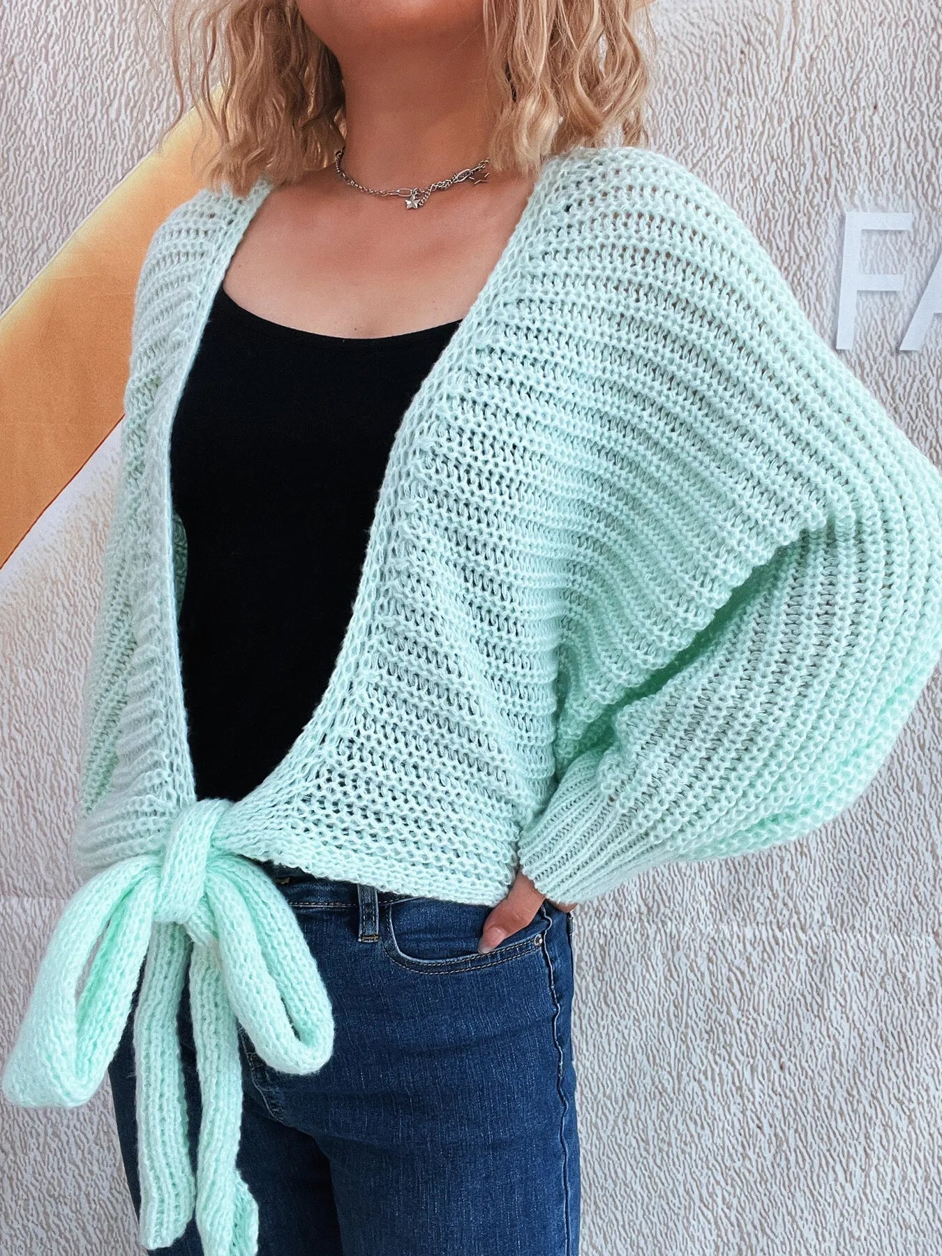 Person wearing a light green knitted cardigan over a black top and blue jeans against a plain background