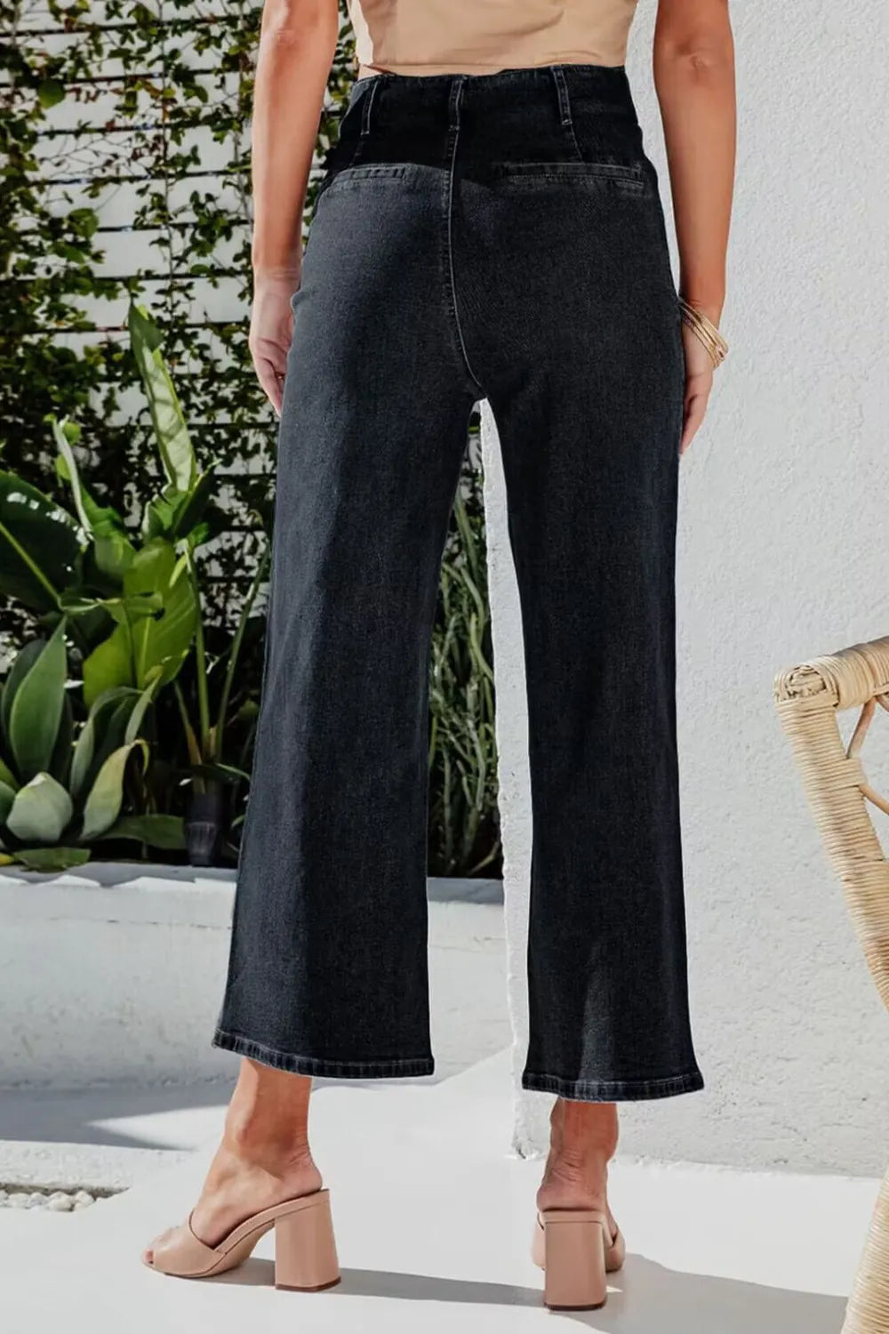 Person wearing black wide-leg jeans standing outdoors with plants and a white wall in the background