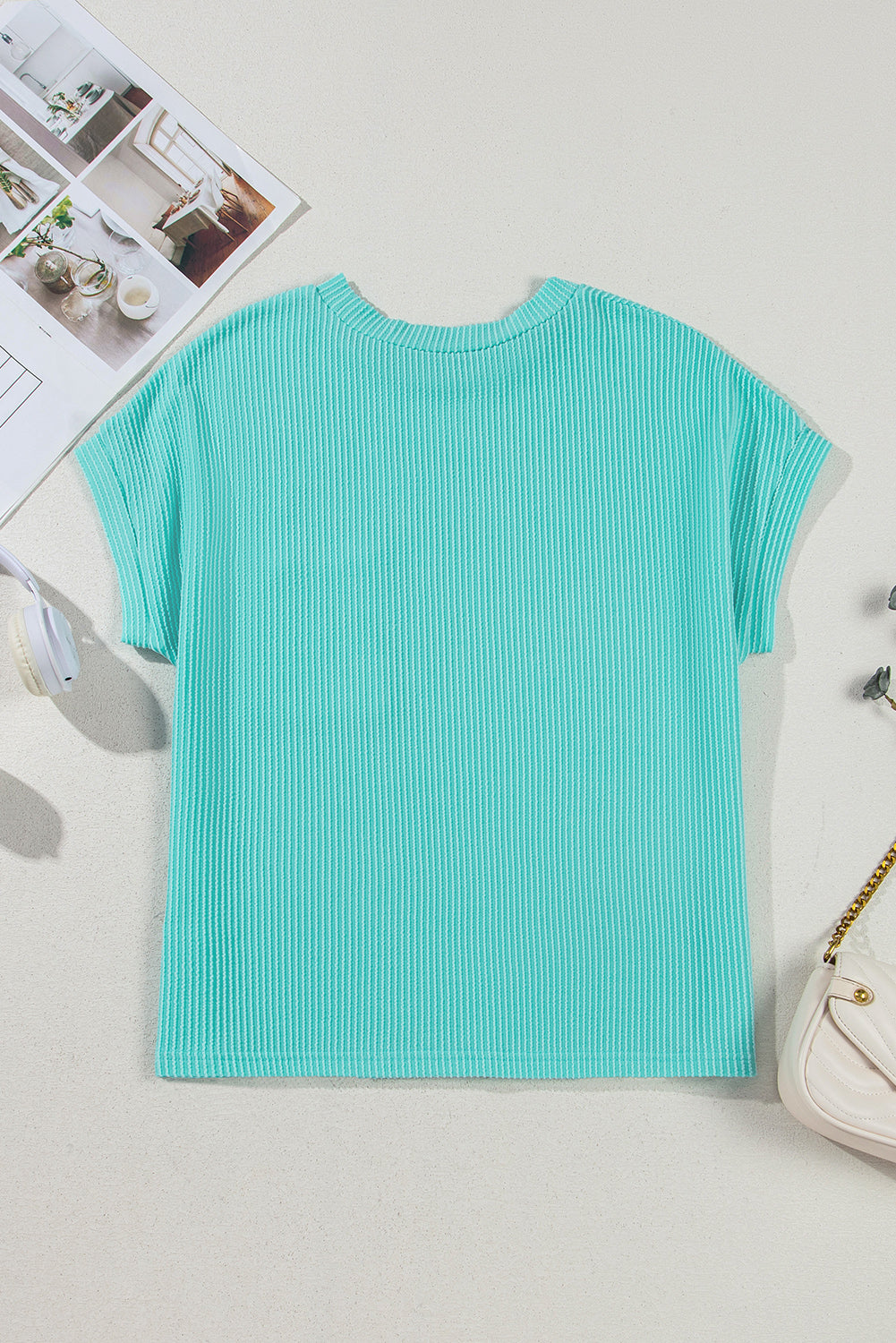 Turquoise ribbed t-shirt on a light gray surface with a magazine and handbag in the background.