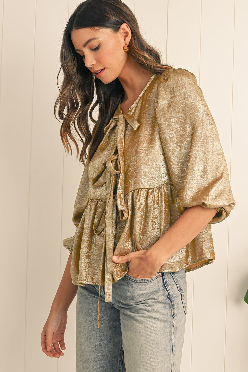 Woman wearing a gold metallic blouse and light blue jeans against a white wall.