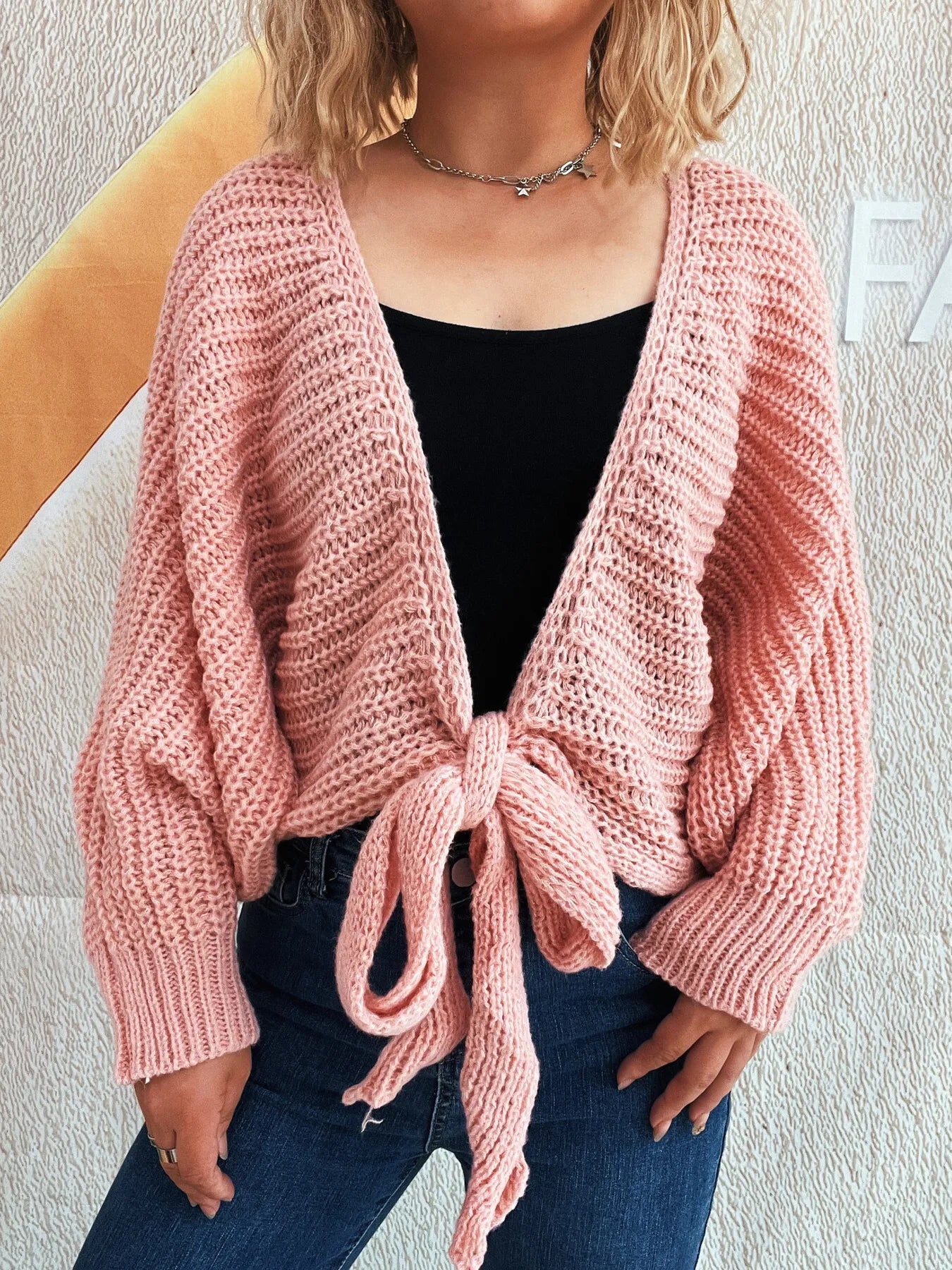 Person wearing a pink knitted cardigan with a tie front, standing against a light-colored wall.