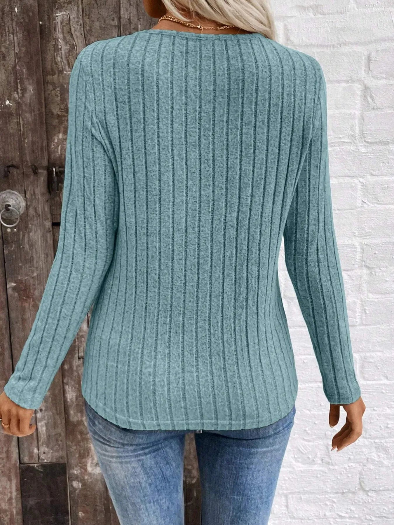 Person wearing a teal ribbed sweater and blue jeans against a wooden and white brick wall.