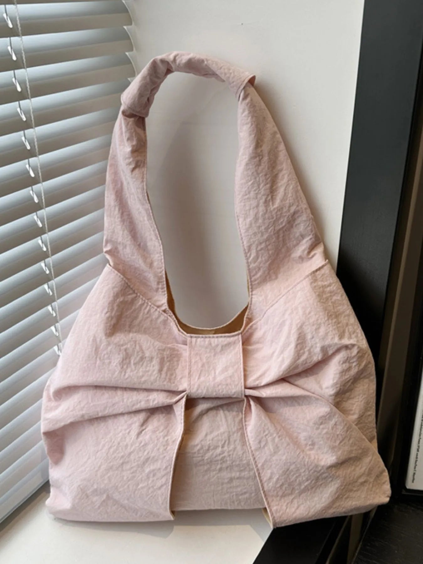 Pink handbag with a bow detail on a white surface next to a window with blinds.