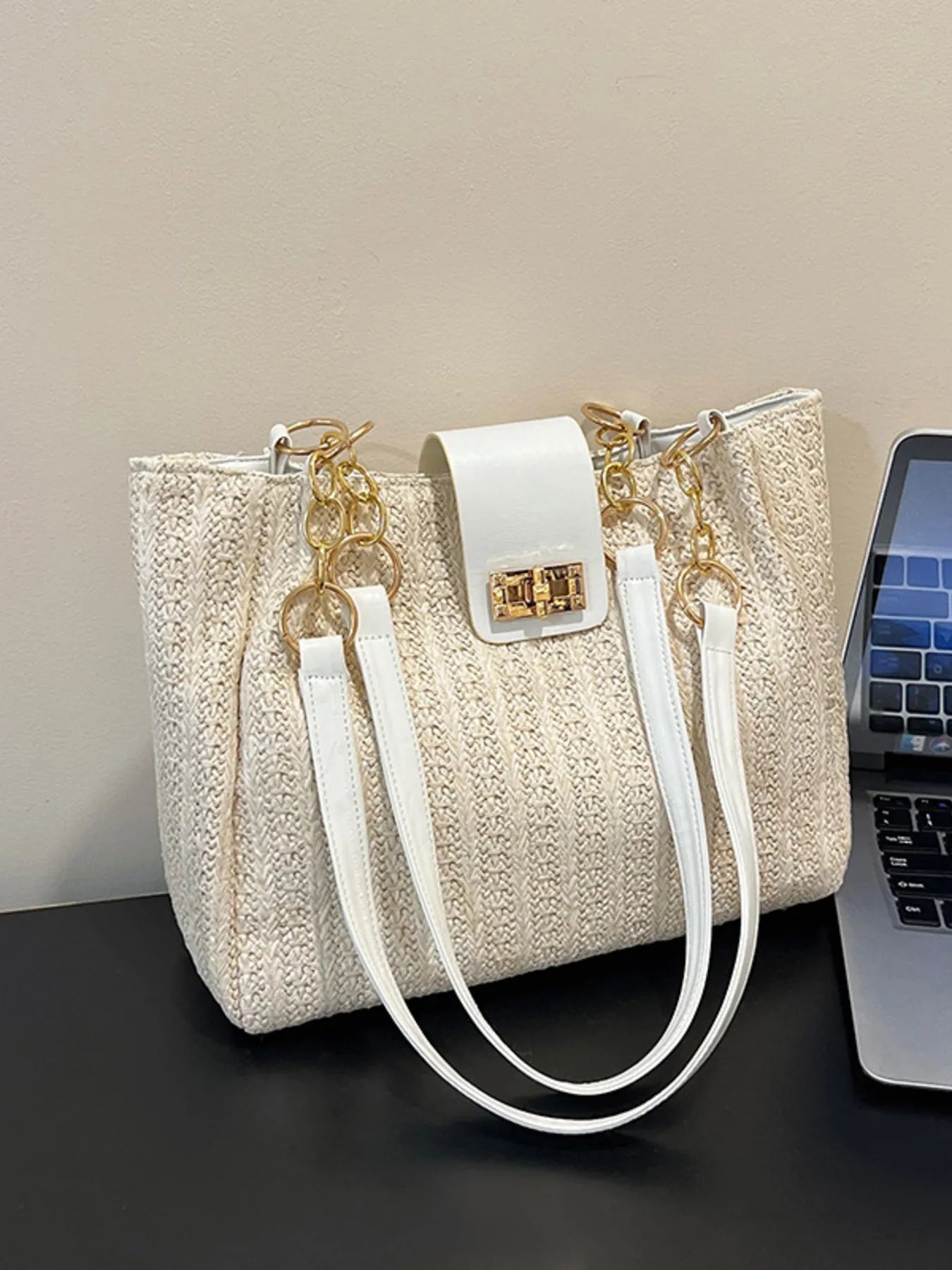 Beige handbag with white straps next to a laptop on a dark surface.