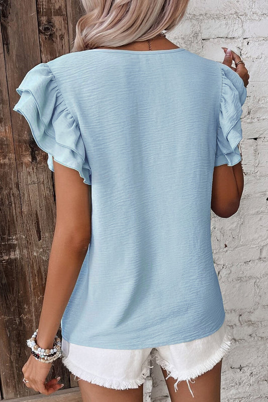 Light blue ruffled sleeve top worn by a person against a wooden and stone wall.