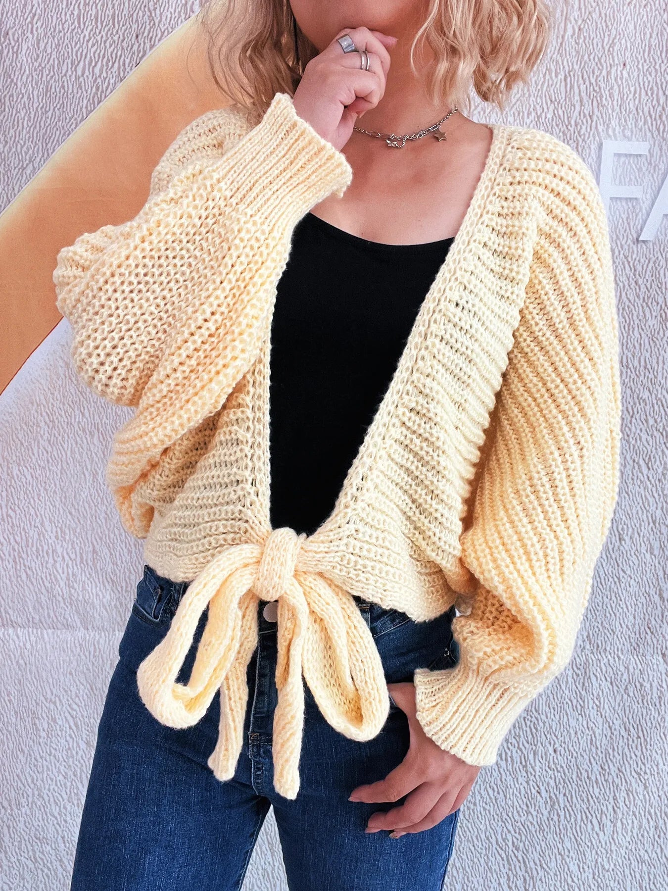 Woman wearing a light yellow knitted cardigan over a black top and blue jeans against a plain background