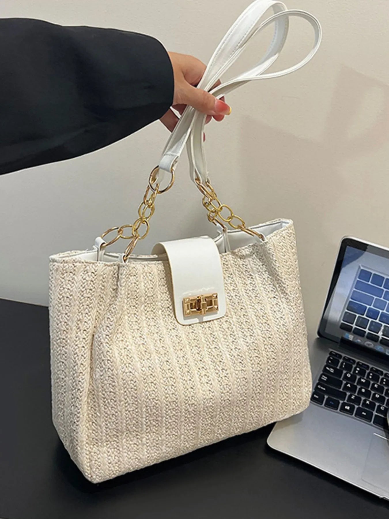 Beige handbag with gold chain handle held by a person, with a laptop in the background.