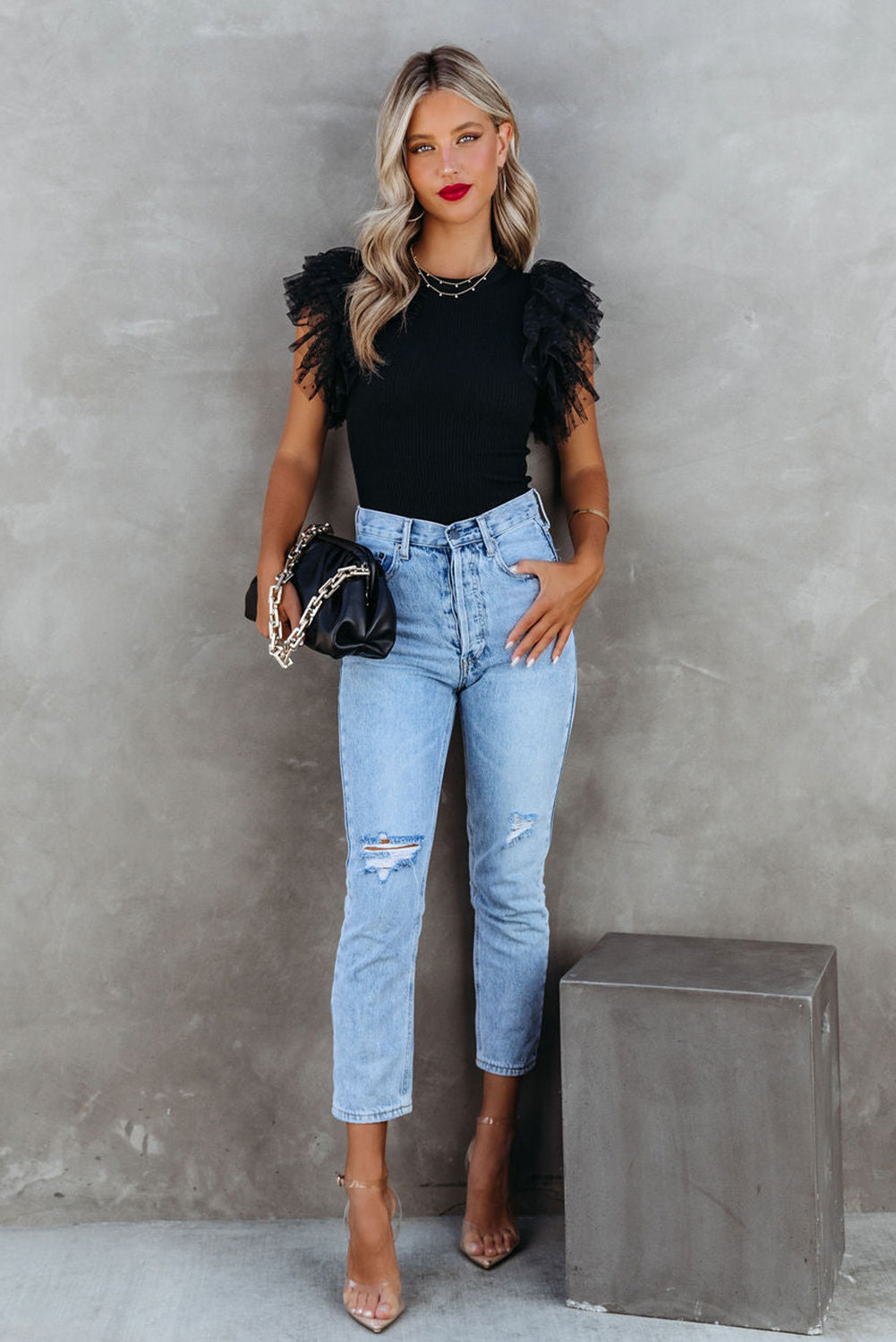 Woman wearing a black top with ruffled sleeves and light blue jeans against a gray background