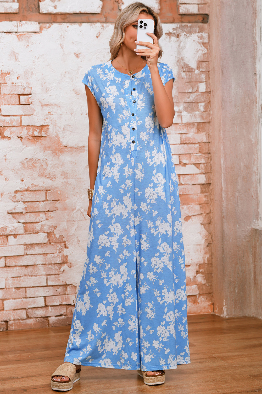 Woman wearing a blue floral dress taking a selfie against a brick wall.