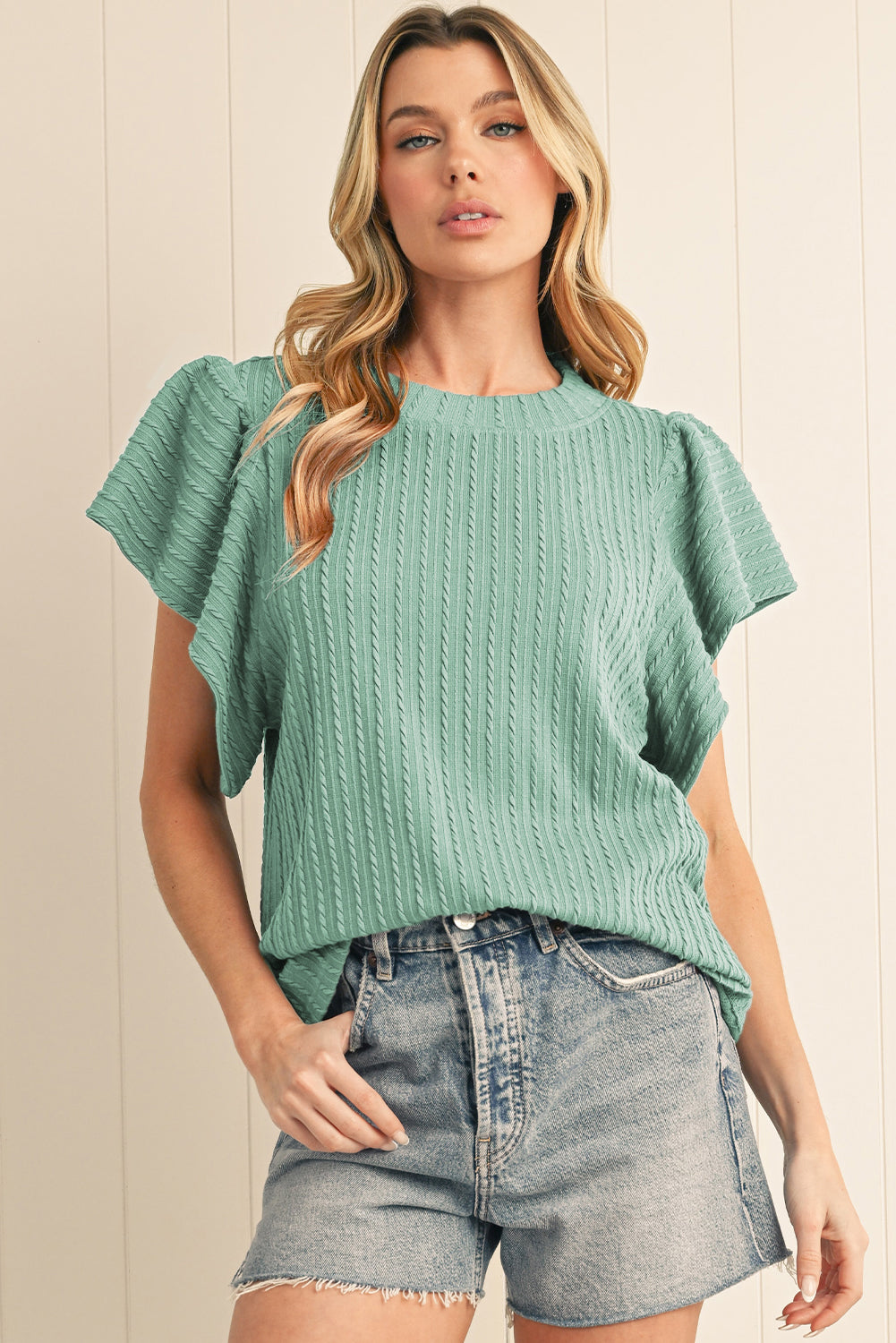Woman wearing a green knitted top and denim shorts against a beige wall.
