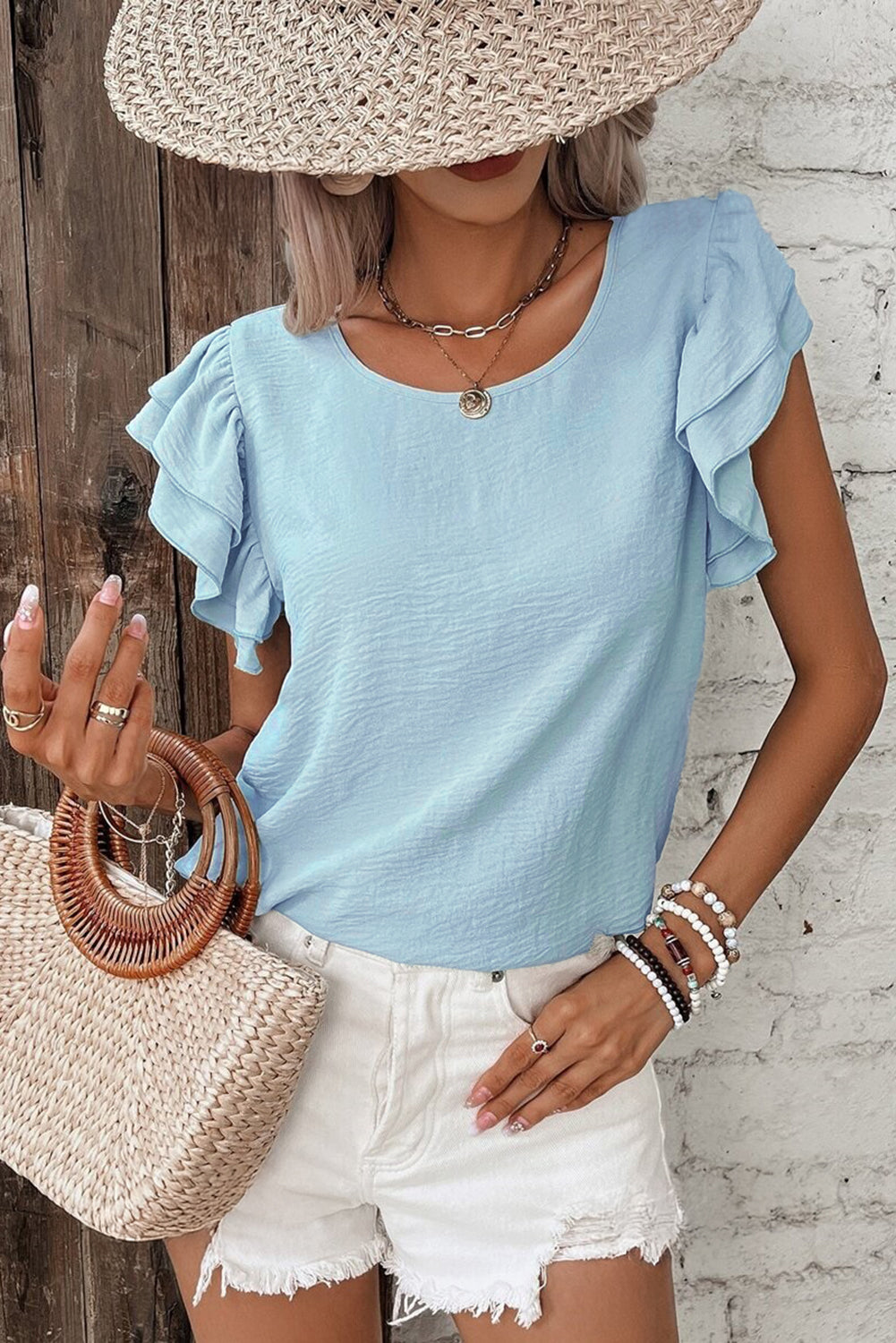 Woman wearing a light blue top, white shorts, and a straw hat against a wooden wall.