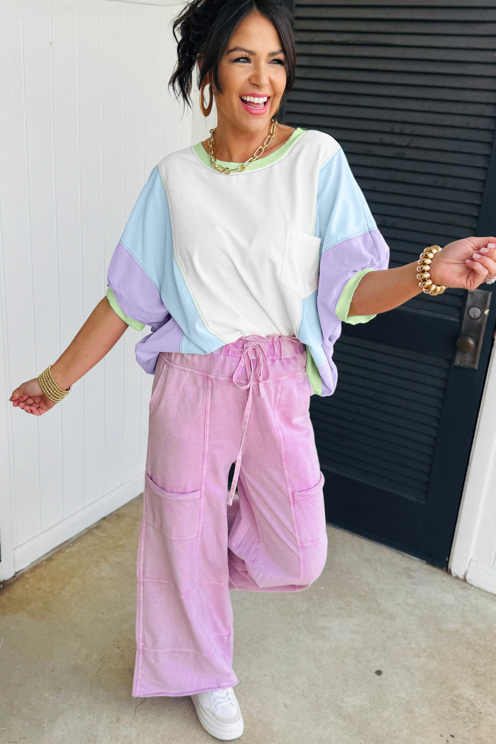 Woman wearing a colorful top and pink pants standing in front of a white wall.