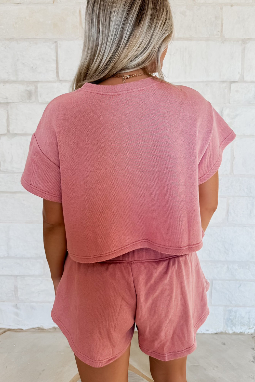 Person wearing a pink crop top and shorts set against a white brick wall.