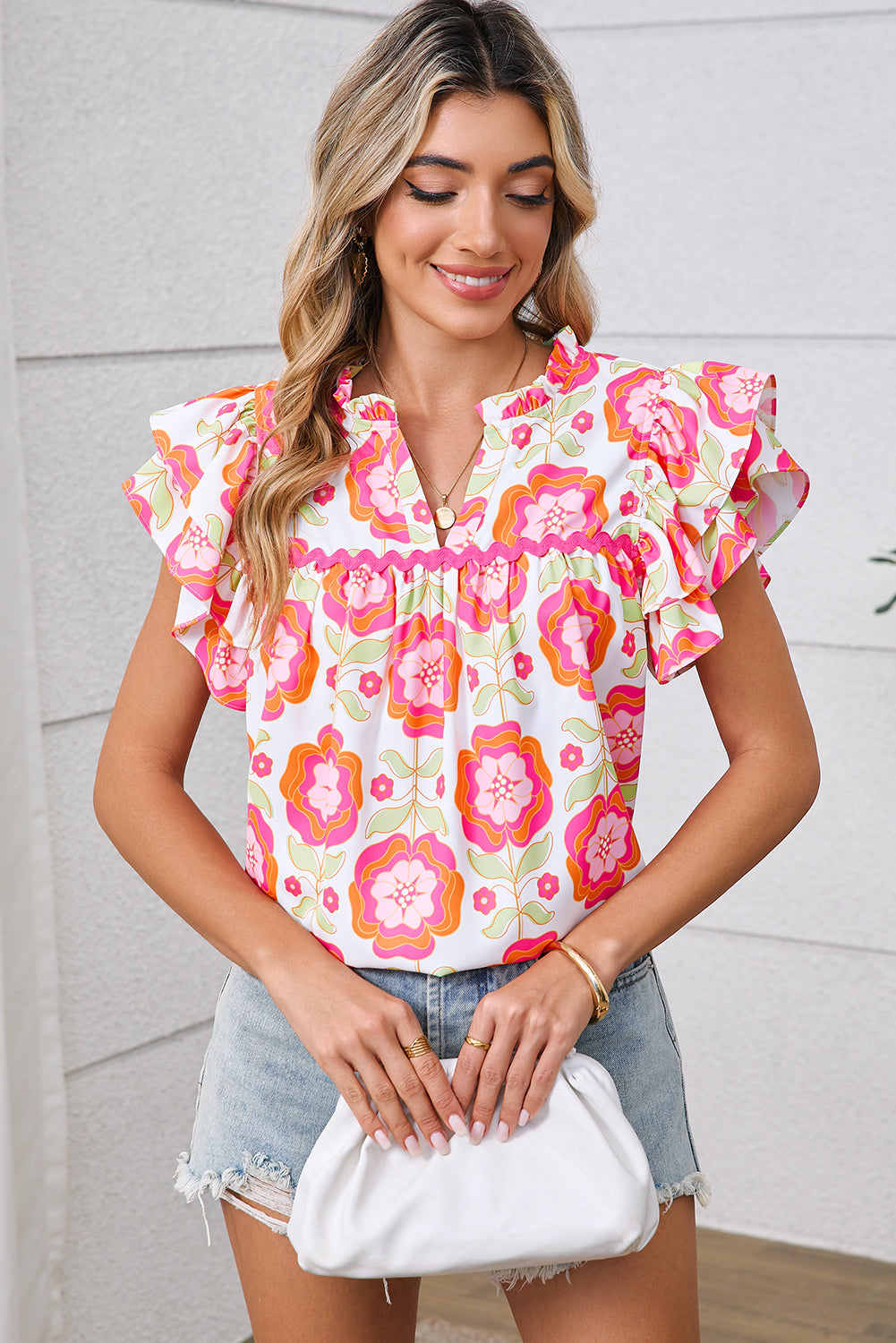 Woman wearing a floral blouse with ruffled sleeves and denim shorts, holding a white clutch.