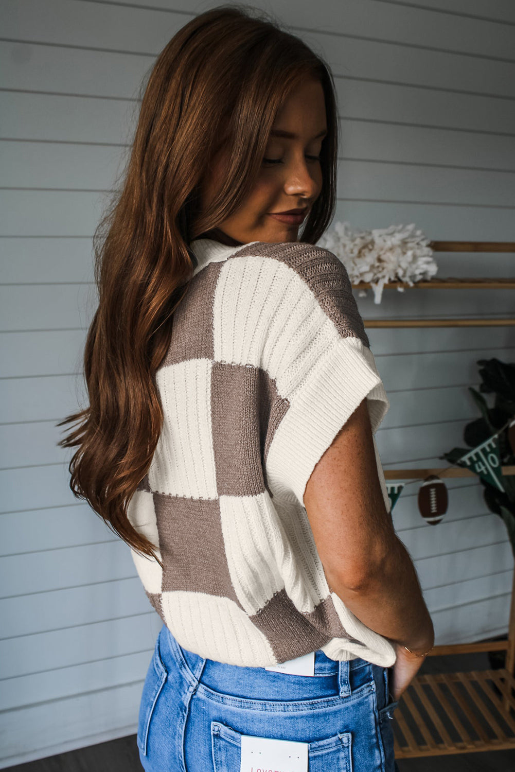 Woman wearing a checkered sweater and blue jeans against a white wall.