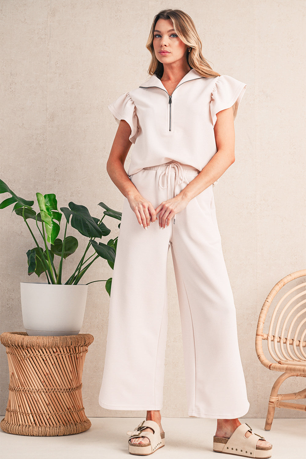 Woman in a white outfit standing in a room with a plant and wicker chair.