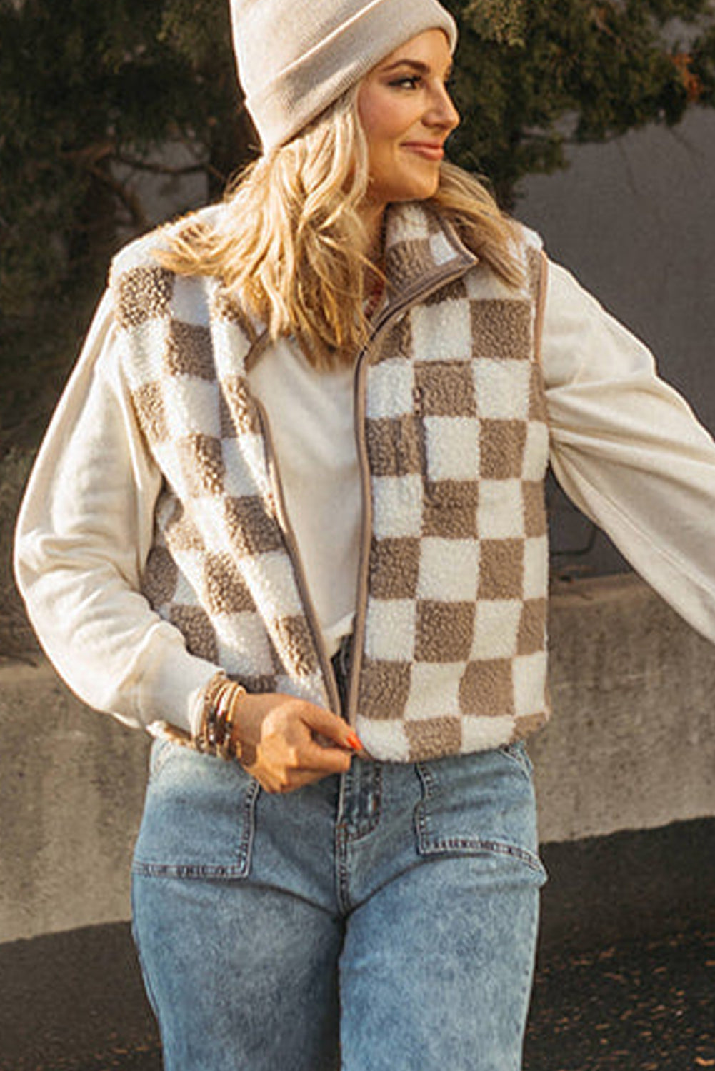 Woman wearing a checkered vest, white sweater, and blue jeans outdoors.