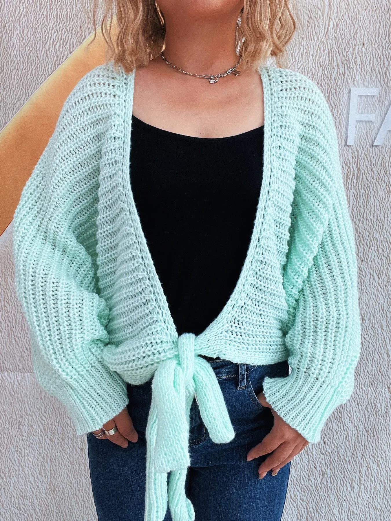 Person wearing a light blue knitted cardigan over a black top and blue jeans against a plain background