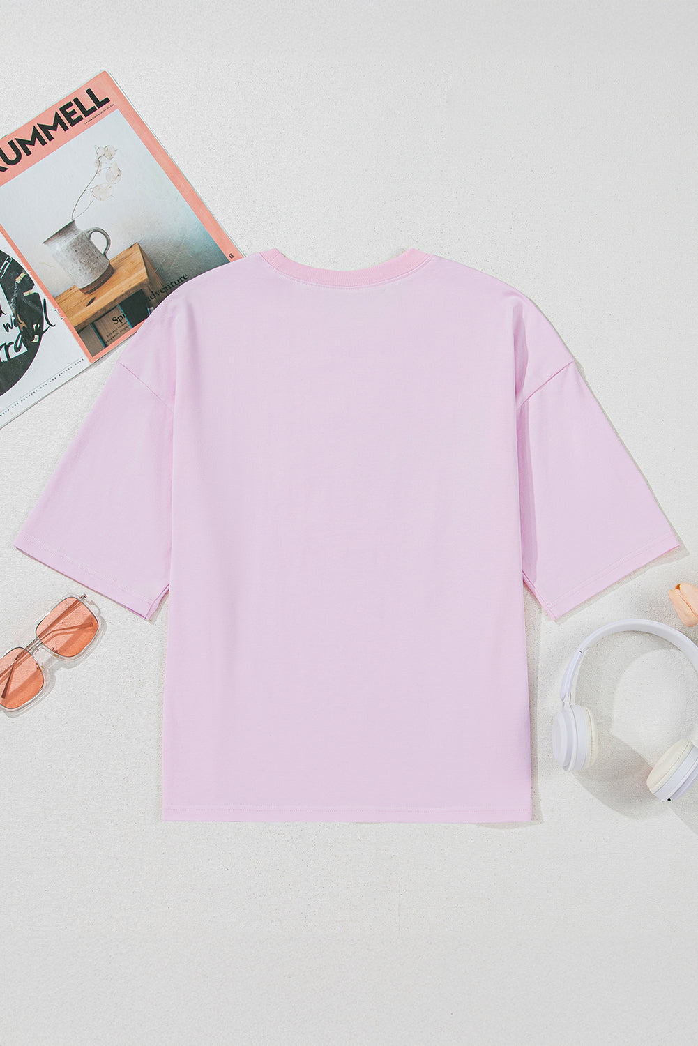 Pink t-shirt on a light gray background with a magazine and sunglasses.