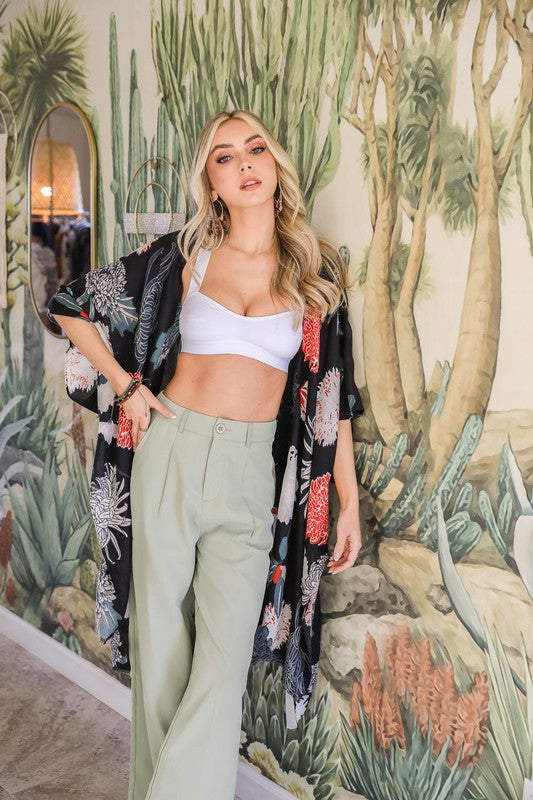 Woman wearing a black floral kimono over a white top and light green pants in front of a nature-themed wall mural.