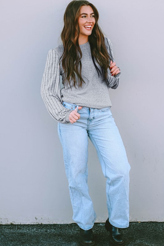 Woman wearing a gray sweater, light blue jeans, and black shoes against a plain background