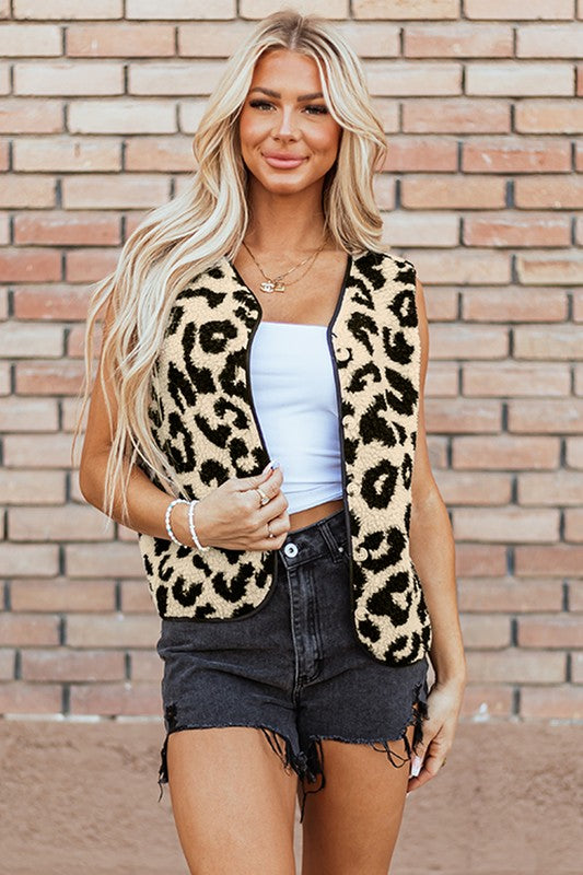 Woman wearing a leopard print vest over a white top and black shorts against a brick wall.
