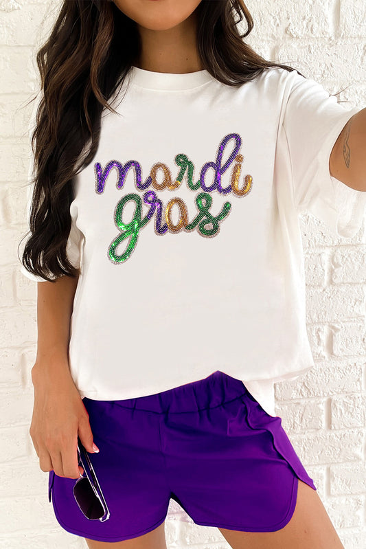 Person wearing a white t-shirt with 'mardi gras' text and purple shorts against a white brick wall.