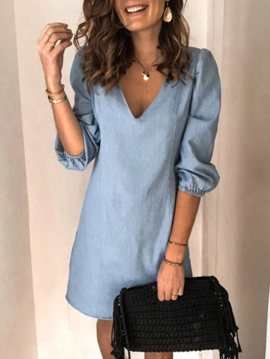 Woman wearing a light blue dress with long sleeves, holding a black clutch against a neutral background.